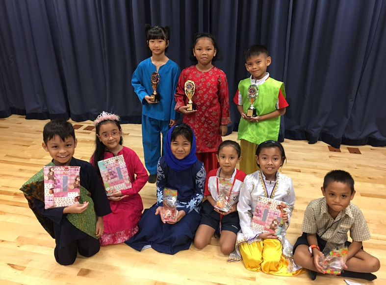 Eight children pose on a wooden floor, some holding trophies and gift bags, dressed in colorful traditional outfits.